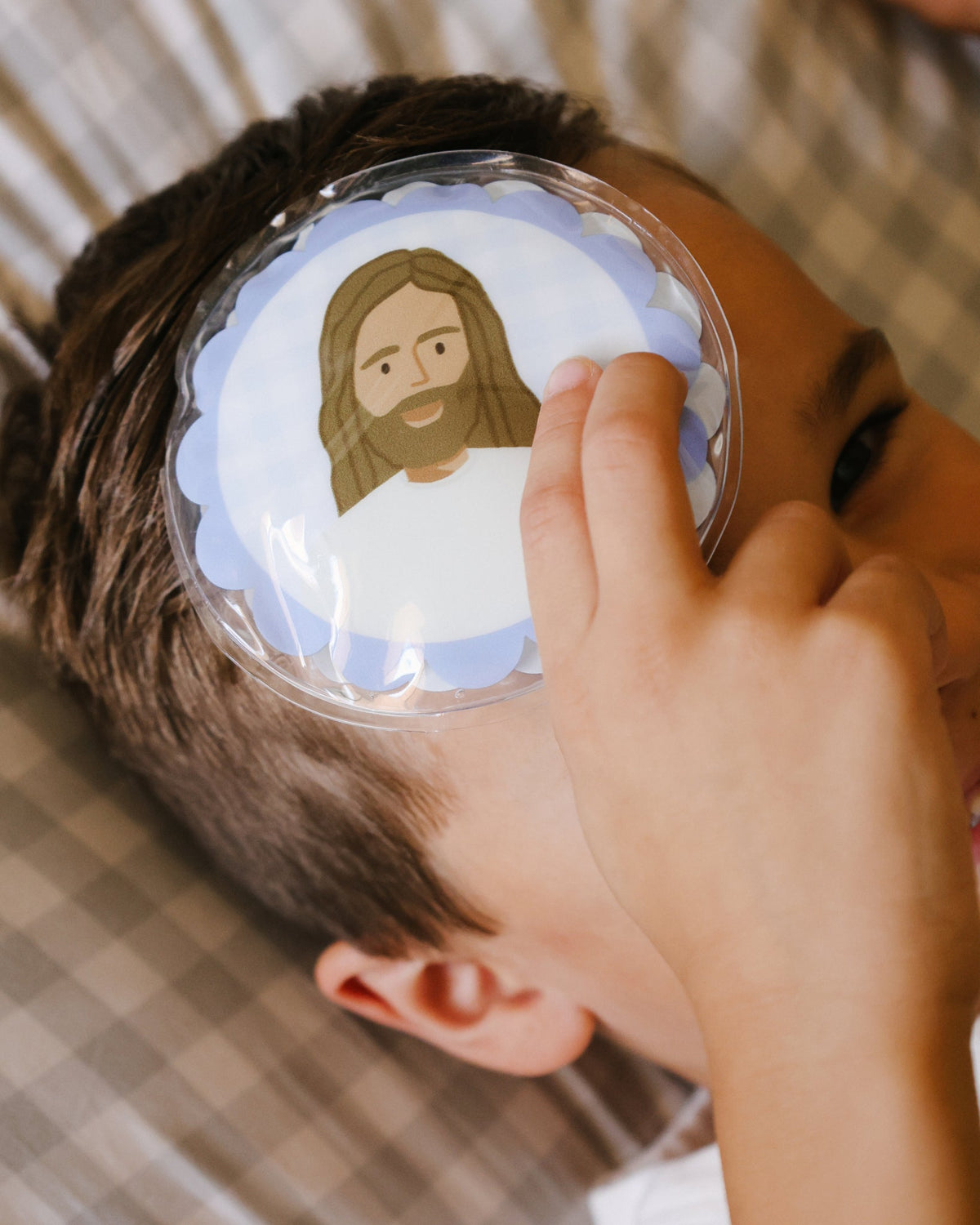 Boy holding a blue Jesus Heals cold pack to his forehead to soothe a boo boo
